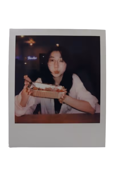 Young woman with glasses holding a takeout container of food, sitting at a table in a dimly lit setting.