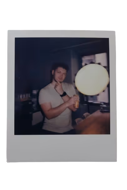 Young man in a light-colored t-shirt holding a canned drink, posing with his hand on his chin in a dimly lit room near a table and window.