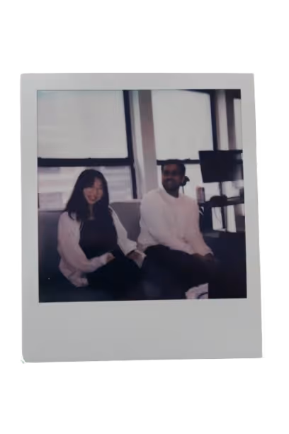 Two people sitting on a couch indoors near large windows, one smiling woman and one man looking to the side.