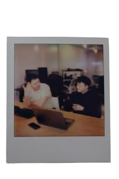 Two people sitting at a desk with a laptop and phone, engaged in conversation in an office setting.