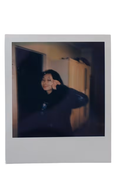 Polaroid photo of a person indoors making a peace sign with their hand near their face, with a dimly lit room and a cabinet in the background.
