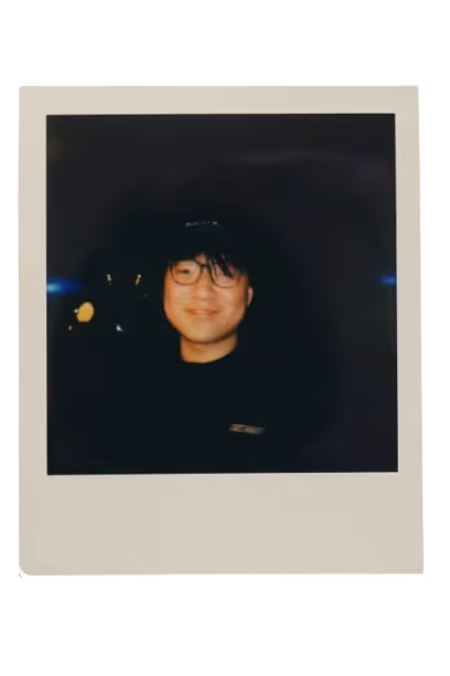 Smiling person wearing glasses and a dark cap in a dimly lit surrounding.