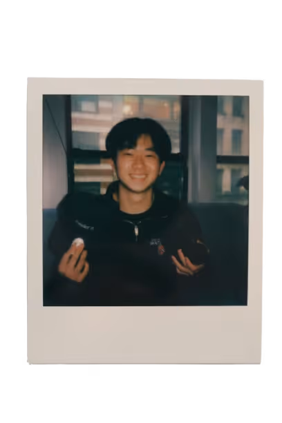 Smiling young man indoors holding a black cap and a dark object with a window and buildings visible behind him.