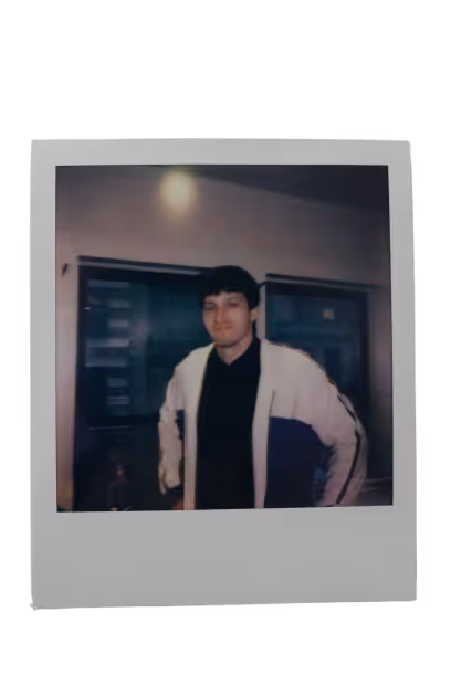 Young man with dark hair wearing a white and blue jacket over a black shirt standing indoors near windows.